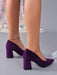 A pair of women's purple velvet block-heel pumps with pointed toes, shown on a model's feet against a gray studio background.