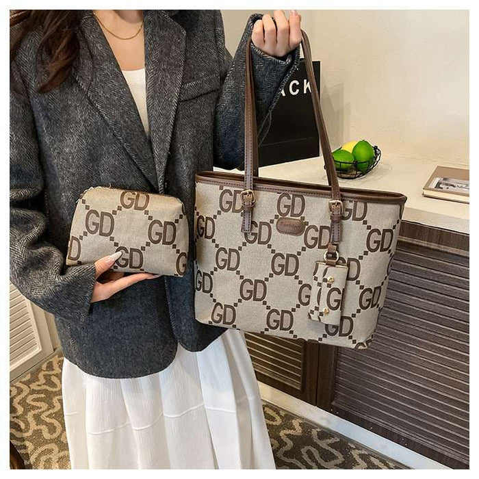 A person wearing a gray blazer and white skirt holds a beige tote bag with brown leather handles and a matching small pouch. The bag has an allover brown 'GD' monogram pattern.