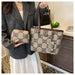 A person wearing a gray blazer and white skirt holds a beige tote bag with brown leather handles and a matching small pouch. The bag has an allover brown 'GD' monogram pattern.