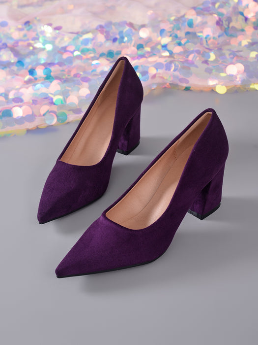 A pair of pointed-toe high heels in deep purple velvet, displayed on a gray surface with a shimmering, iridescent sequin backdrop.