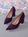 A pair of pointed-toe high heels in deep purple velvet, displayed on a gray surface with a shimmering, iridescent sequin backdrop.