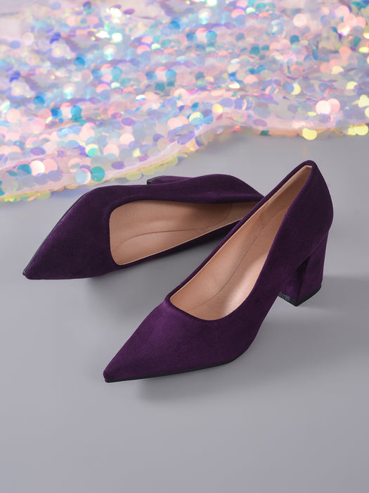 A pair of deep purple velvet pointed-toe pumps with block heels, displayed on a gray surface with a shimmering, iridescent sequin fabric in the background.