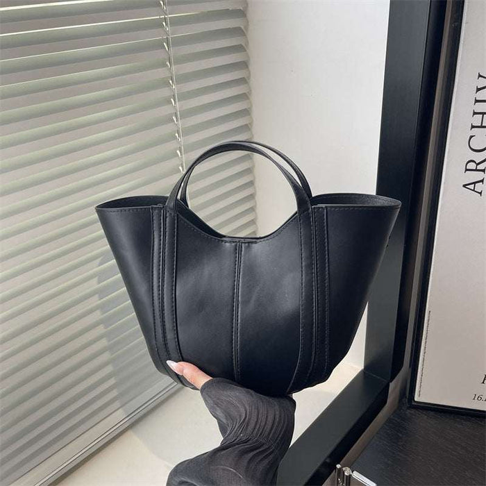 A black leather tote bag with dual top handles, held by a hand wearing a dark gray knit sleeve. The bag is positioned in front of a window with white blinds, next to a black frame and a book.