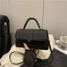 A black faux-leather handbag with a top handle and shoulder strap, held by a hand wearing a sheer black sleeve. The handbag is placed on a white textured blanket with a black suitcase and a white cup in the background.