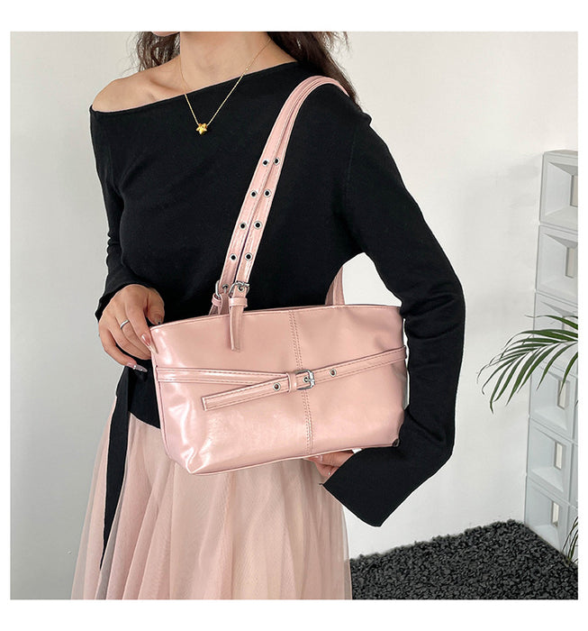 A woman wearing a black off-the-shoulder long-sleeve top and a light pink pleated skirt carries a shiny metallic pink shoulder bag with a wide strap and silver studs.