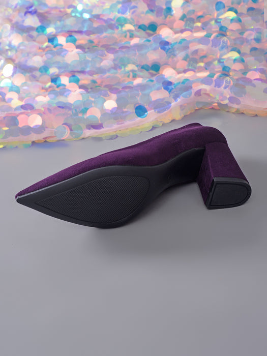A single purple velvet pointed-toe high heel with a black sole and block heel, photographed on a gray surface with a shimmering sequin backdrop.