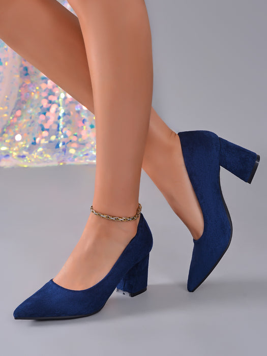 A pair of navy blue suede block-heel pumps with a pointed toe, shown on a model's feet against a gray background.