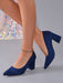 A pair of navy blue suede block-heel pumps with a pointed toe, shown on a model's feet against a gray background.