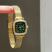 Gold wristwatch with green face held by a hand against a gray background