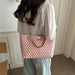 A woman viewed from behind wearing a light pink and beige patterned tote bag with a gold chain strap, styled with a white blouse and light blue jeans in an indoor setting.