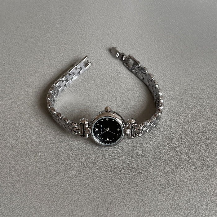 A silver-tone metal bracelet watch with a round black dial, photographed on a light gray textured surface.