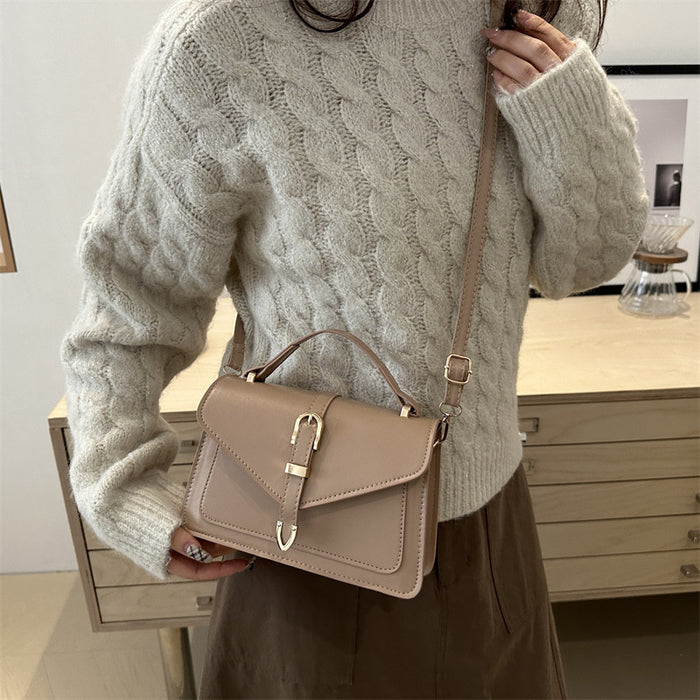 A woman wearing a cream cable-knit sweater and brown skirt carries a taupe leather crossbody handbag with a top handle and buckle closure.