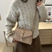 A woman wearing a cream cable-knit sweater and brown skirt carries a taupe leather crossbody handbag with a top handle and buckle closure.