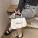 A person seated on a wooden bench holds a small structured handbag. The bag is off-white/cream with gold-tone hardware and a circular white charm printed with the word FASHION.