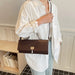 A person wearing an oversized white button-up shirt and light blue jeans carries a dark brown woven crossbody bag with a gold clasp in an indoor setting.