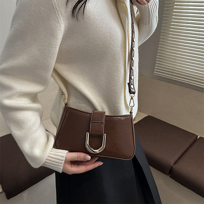 A person wearing a cream knit cardigan and black skirt carries a small brown leather shoulder bag with a silver-tone U-shaped metal detail and a chain strap in an indoor setting.