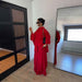 A woman wearing a bright red two-piece outfit with a high-neck long-sleeve top and matching wide-leg skirt, standing in a minimalist room with a large floor mirror and a black wardrobe door.
