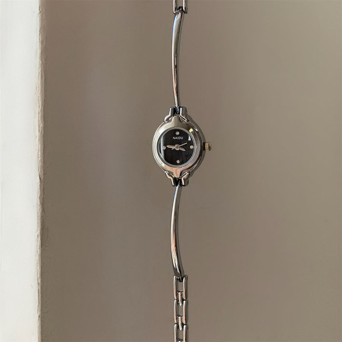 A silver-tone metal link watch with a round black dial and small crystal hour markers, hanging against a plain beige wall.