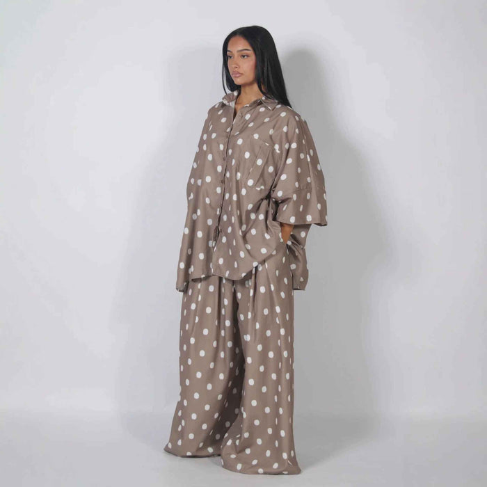 A woman modeling a matching lounge set consisting of a long-sleeve button-up shirt and wide-leg pants, both in a taupe-brown color with a white polka-dot pattern, set against a plain light gray studio background.