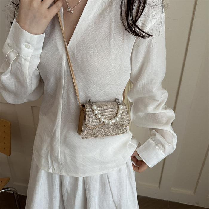 A woman wearing a white linen long-sleeve blouse and white skirt carries a small woven clutch bag with a pearl-embellished flap and a beige shoulder strap in an indoor setting.