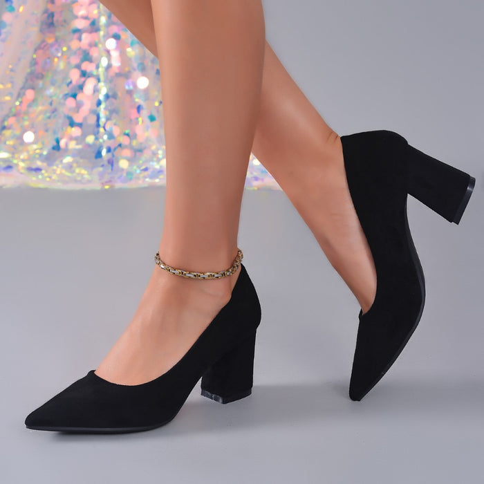 Black pointed-toe block-heel pumps worn on a model's feet, featuring a gold chain anklet on one shoe, set against a gray background with a sequined skirt visible in the upper left.