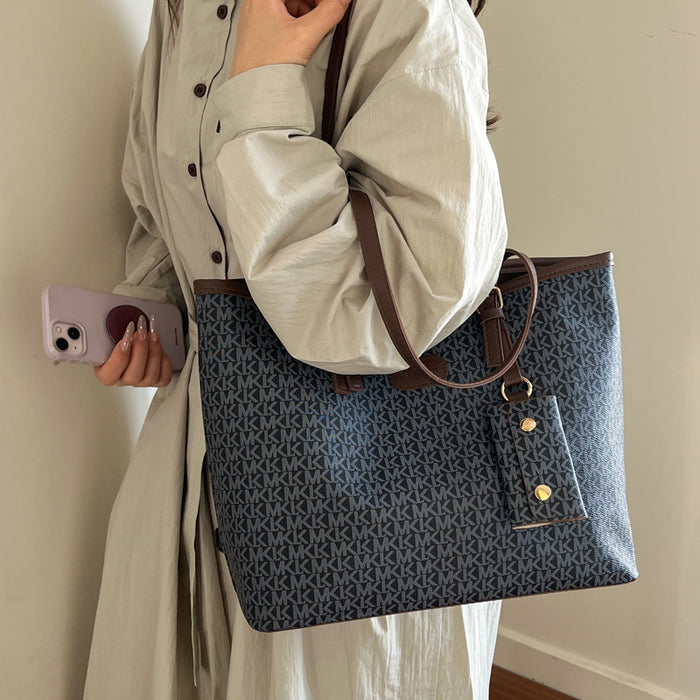 A person wearing a light beige long-sleeve shirt carries a large blue tote bag with an allover monogram pattern and brown shoulder straps. The person is holding a light purple smartphone against a plain indoor wall.