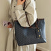A person wearing a light beige long-sleeve shirt carries a large blue tote bag with an allover monogram pattern and brown shoulder straps. The person is holding a light purple smartphone against a plain indoor wall.