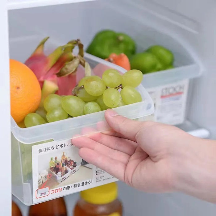 A hand holding a clear plastic refrigerator organizer bin filled with green grapes, an orange, a dragon fruit, and other fruits inside a refrigerator.