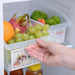 A hand holding a clear plastic refrigerator organizer bin filled with green grapes, an orange, a dragon fruit, and other fruits inside a refrigerator.