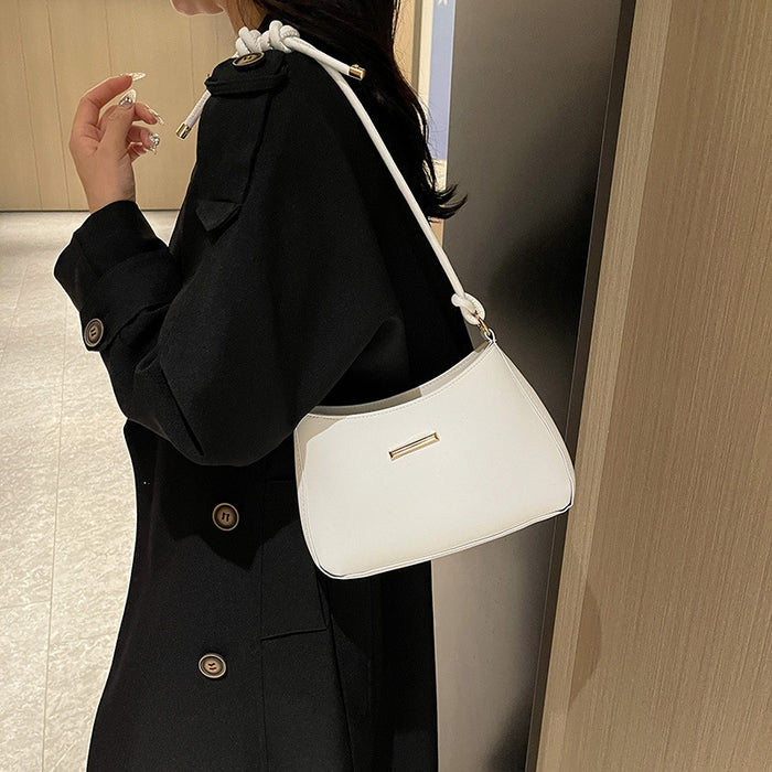 A woman wearing a black coat carries a white crescent-shaped shoulder bag with a knotted strap detail and a small gold metal accent on the front, photographed indoors against a neutral wall and wood paneling.