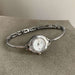 A silver-tone bangle watch with a round white dial and a small visible 'MOON' brand mark, resting on a textured gray surface.