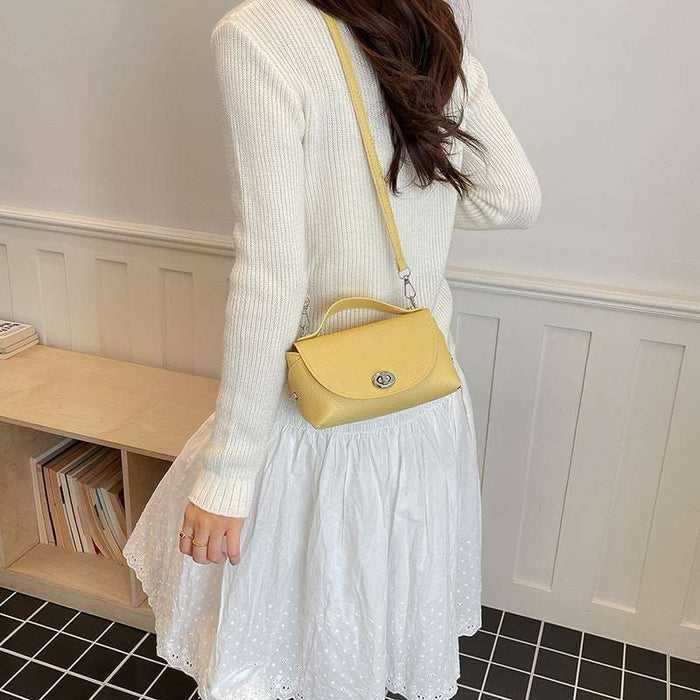 A woman wearing a white knit sweater and white skirt carries a small yellow crossbody handbag with a top handle and silver clasp in an indoor room with white paneled walls and a wooden shelf.