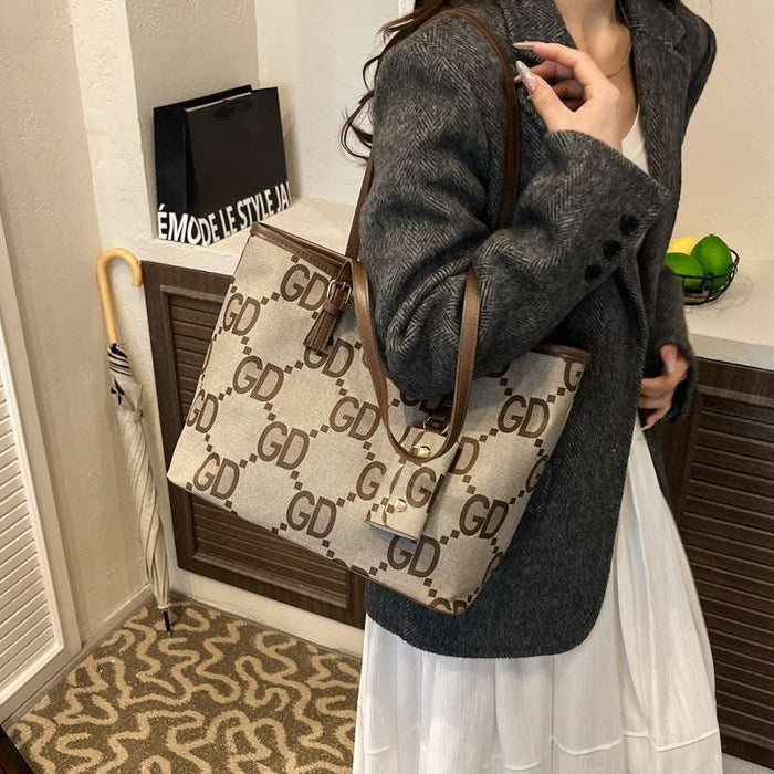 A woman wearing a gray blazer and white skirt carries a large beige tote bag with a brown monogram pattern and brown leather straps in an indoor setting.