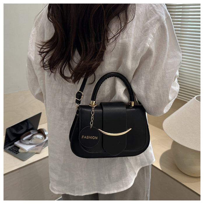 A woman wearing a white linen shirt carries a black structured handbag with gold-tone hardware and a hanging circular charm that reads 'FASHION'. The photo is taken from behind in an indoor setting with a desk, laptop, and lamp visible in the background.