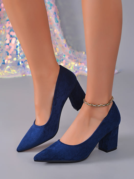 A pair of navy blue suede block-heel pumps with pointed toes, shown on a model's feet against a gray studio background with a sequined dress visible in the background.
