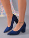 A pair of navy blue suede block-heel pumps with pointed toes, shown on a model's feet against a gray studio background with a sequined dress visible in the background.