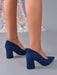 A pair of women's navy blue suede block-heel pumps with pointed toes, shown on a model's feet against a gray studio background with a sparkly fabric backdrop.