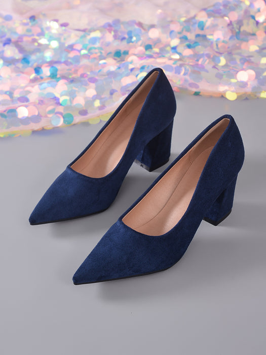 A pair of navy blue suede high-heeled pumps with pointed toes and block heels, displayed on a gray surface with a colorful, glittery sequin backdrop.