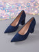 A pair of navy blue suede high-heeled pumps with pointed toes and block heels, displayed on a gray surface with a colorful, glittery sequin backdrop.