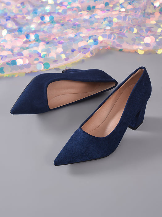 A pair of navy blue suede pointed-toe pumps with a low block heel, displayed on a gray surface with a sparkling, iridescent sequin fabric in the background.