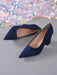 A pair of navy blue suede pointed-toe pumps with a low block heel, displayed on a gray surface with a sparkling, iridescent sequin fabric in the background.