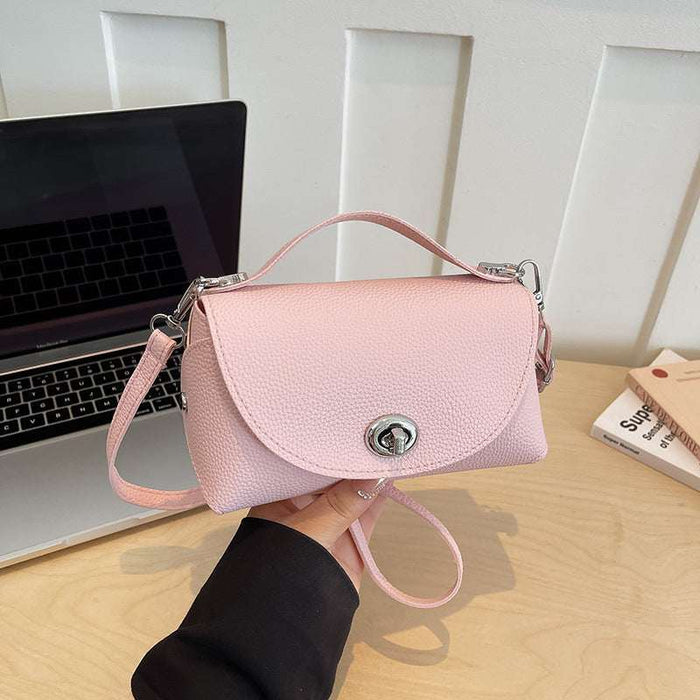 A light pink textured leather handbag with a top handle and detachable shoulder strap, held by a hand over a light wood desk with a laptop in the background.