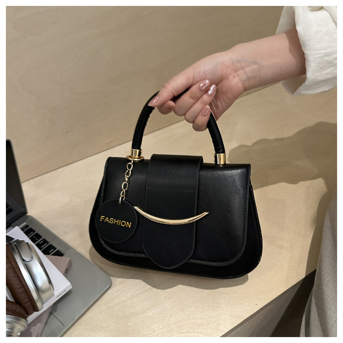 A black faux-leather handbag with gold-tone hardware, held by a person's hand on a light wood desk. The bag features a top handle, a front flap with a central strap, a curved gold-tone metal bar, and a round black tag with the word 'FASHION' in gold.