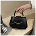 A black faux-leather handbag with gold-tone hardware, held by a person's hand on a light wood desk. The bag features a top handle, a front flap with a central strap, a curved gold-tone metal bar, and a round black tag with the word 'FASHION' in gold.