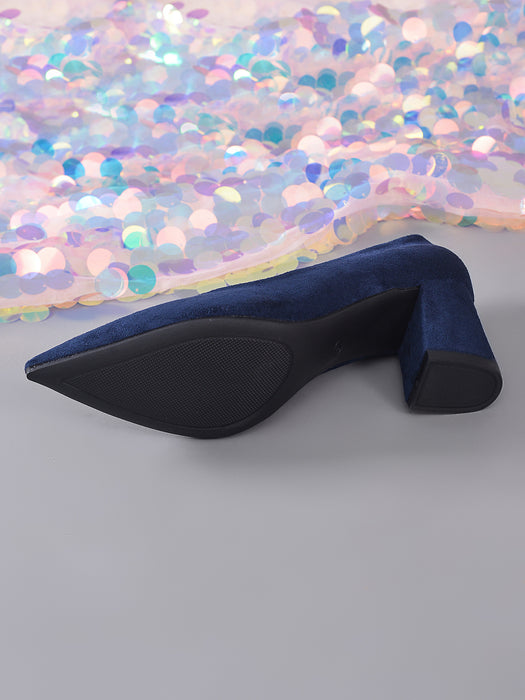 A single navy blue velvet pointed-toe heeled boot shown from a side angle, with a black textured outsole and block heel, set against a gray background with a shimmering sequin fabric backdrop.