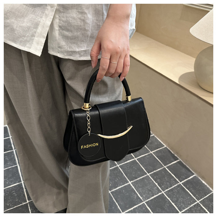 A person wearing a light gray linen shirt and taupe trousers holds a structured black handbag with gold-tone hardware and a circular hanging charm that reads FASHION. The setting appears to be a neutral indoor space with a tiled floor and beige wall.