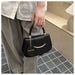 A person wearing a light gray linen shirt and taupe trousers holds a structured black handbag with gold-tone hardware and a circular hanging charm that reads FASHION. The setting appears to be a neutral indoor space with a tiled floor and beige wall.
