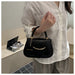 A person wearing a white linen shirt and beige pants holds a small black handbag with gold-tone hardware and a circular charm that reads 'FASHION'. The setting appears to be a softly lit indoor room with a desk and lamp in the background.