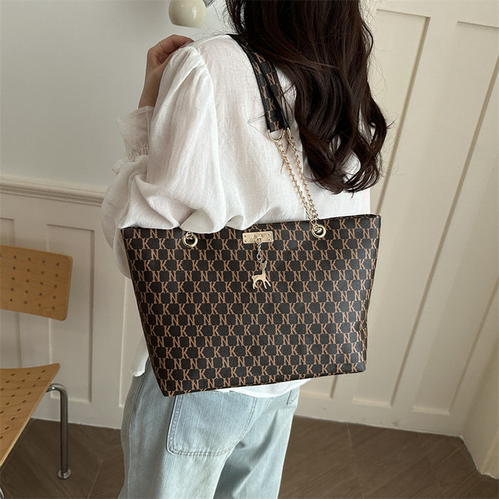 A woman wearing a white blouse and light blue jeans carries a large brown tote bag with a repeating gold monogram pattern, a gold chain strap, and a small hanging charm, photographed indoors against a white paneled wall.