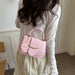 A woman wearing a cream-colored long-sleeve top and skirt carries a light pink faux-leather handbag with a top handle and gold-tone hardware. The handbag features a round hanging charm with the word 'FASHION' printed on it.